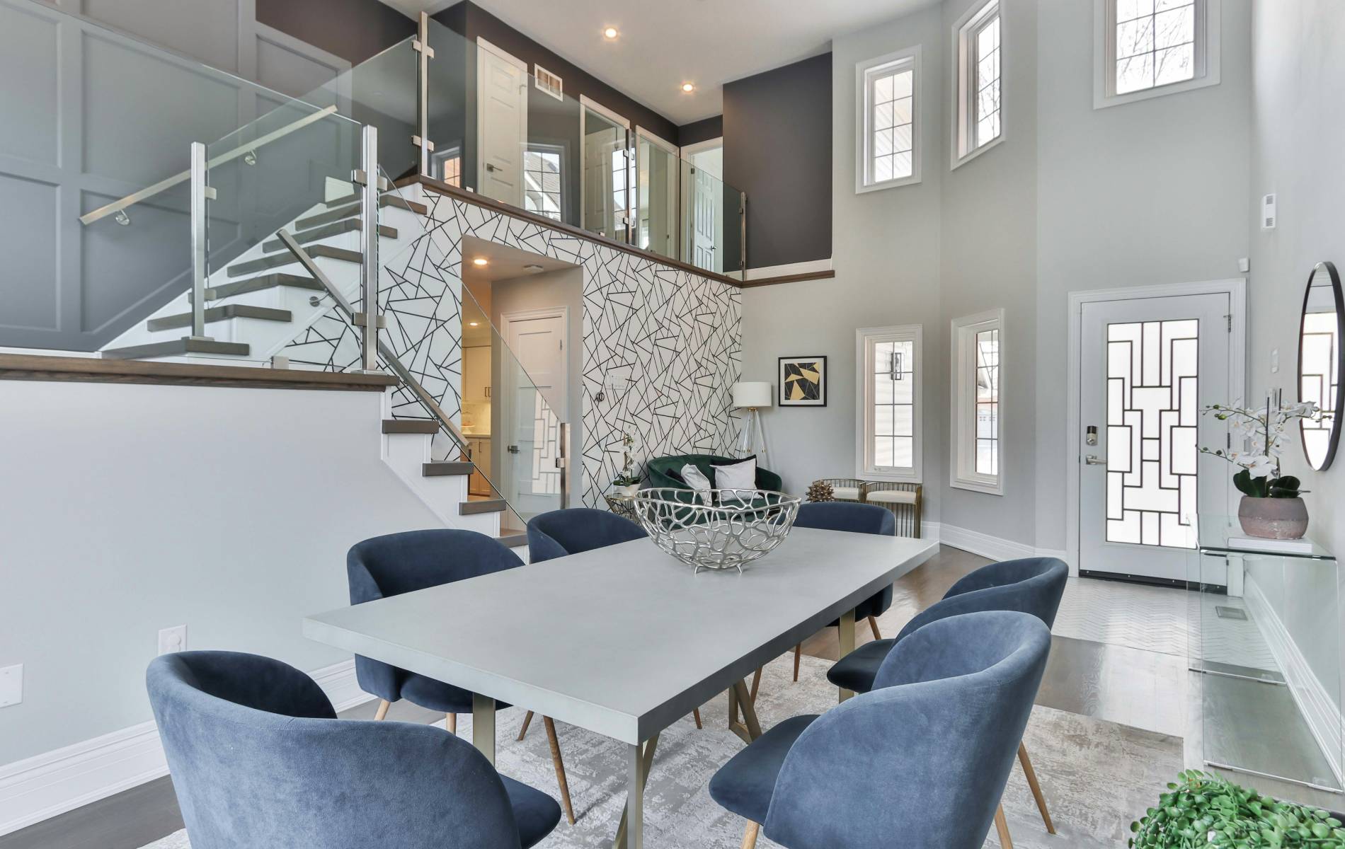 Comedor de diseño moderno con mesa rectangular clara, sillas tapizadas en azul y decoración en tonos neutros. Escalera con barandilla de cristal, pared con motivos geométricos y planta superior con pasillo acristalado.