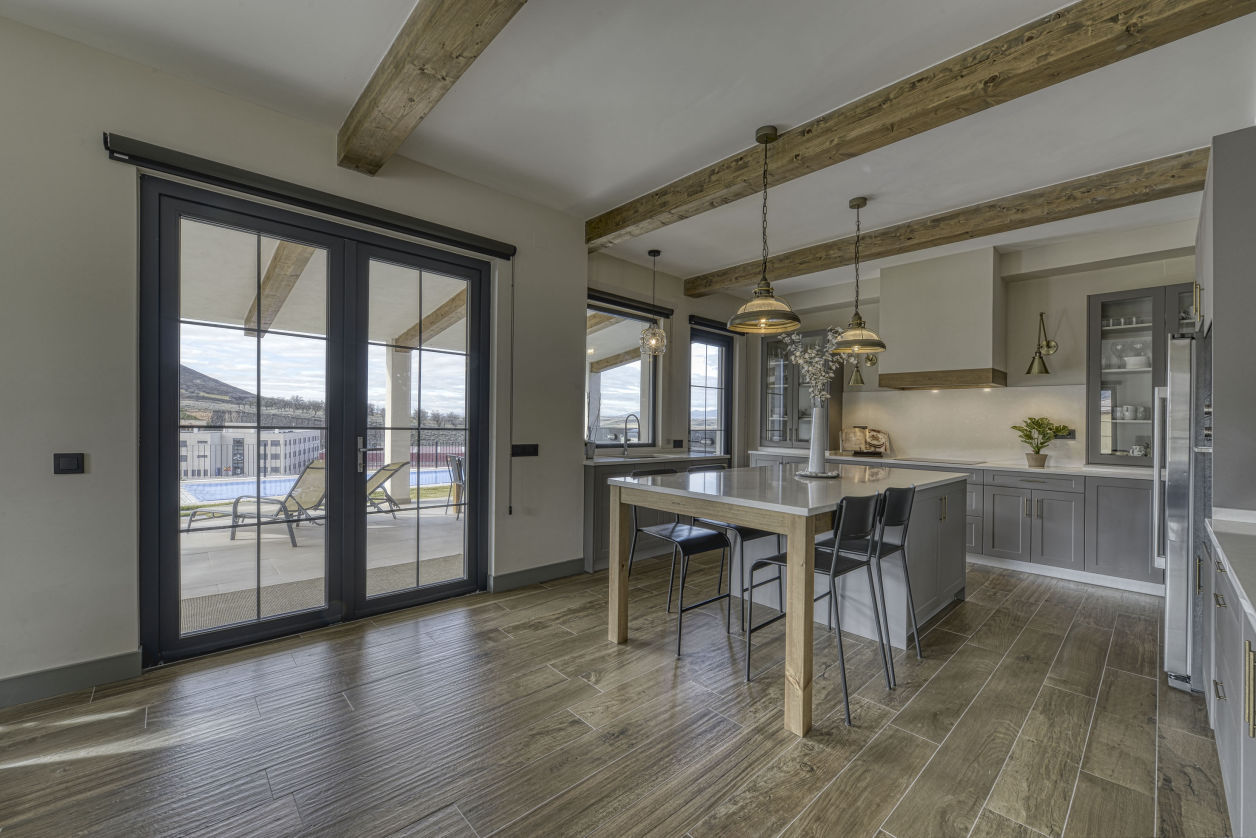 Cocina abierta con isla central, vigas de madera vista en el techo y grandes puertas acristaladas con perfilería oscura que dan acceso a una terraza con vistas. Espacio luminoso, funcional y de estilo contemporáneo con toques rústicos.