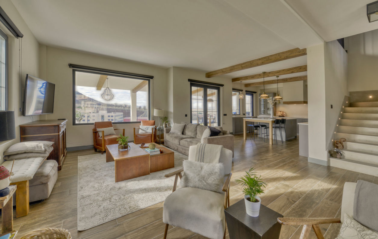 Salón amplio y luminoso con zona de estar, comedor y cocina integrada, suelos de madera, techos con vigas vistas y grandes ventanales con perfilería oscura. Espacio moderno con toques cálidos y acceso a la planta superior por escalera lateral.